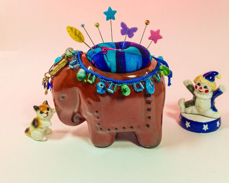 May include: A brown ceramic elephant pincushion with a blue and purple fabric pincushion on top. The elephant is decorated with colourful beads and a gold chain. A small porcelain clown figurine is on a blue and white base to the right of the elephant.