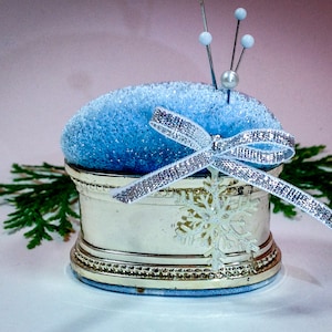 Könnte beinhalten: Ein dekoratives Nadelkissen mit hellblauem, glitzerndem Oberteil und silberner Basis. Ein silbernes Band und eine Schneeflocke akzentuieren das Design. Drei Nadeln mit weißen Kugeln sind in das Kissen gesteckt. Grünes Laub im Hintergrund.
