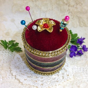 May include: A red velvet pincushion with a gold flower design and colorful pins. The pincushion is in a round, multi-colored container with a gold trim.