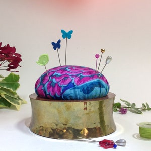 May include: A brass pincushion with a round, floral patterned fabric top. The pincushion has several colorful pins and a green bird pin stuck in it.