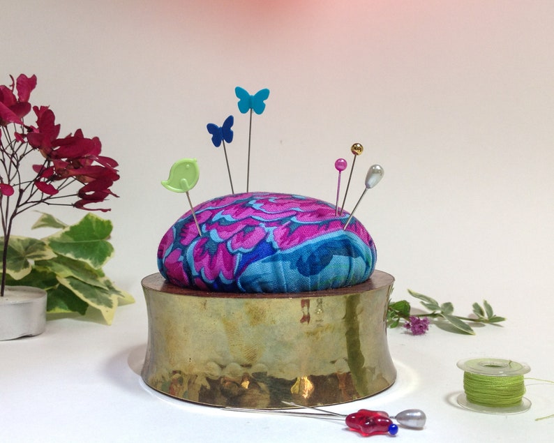 May include: A brass pincushion with a round, floral patterned fabric top. The pincushion has several colorful pins and a green bird pin stuck in it.