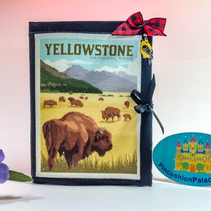 May include: A black fabric journal with a vintage Yellowstone National Park design featuring a brown bison and a mountain landscape. The journal has a black ribbon closure and a yellow bird charm with a red and black plaid bow.