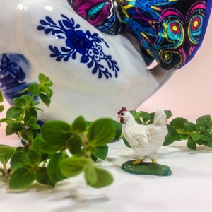 May include: A white ceramic rooster figurine with blue and white painted details, a colourful fabric scarf, and a small white chicken figurine with red accents. The figurines are surrounded by green leafy plants.