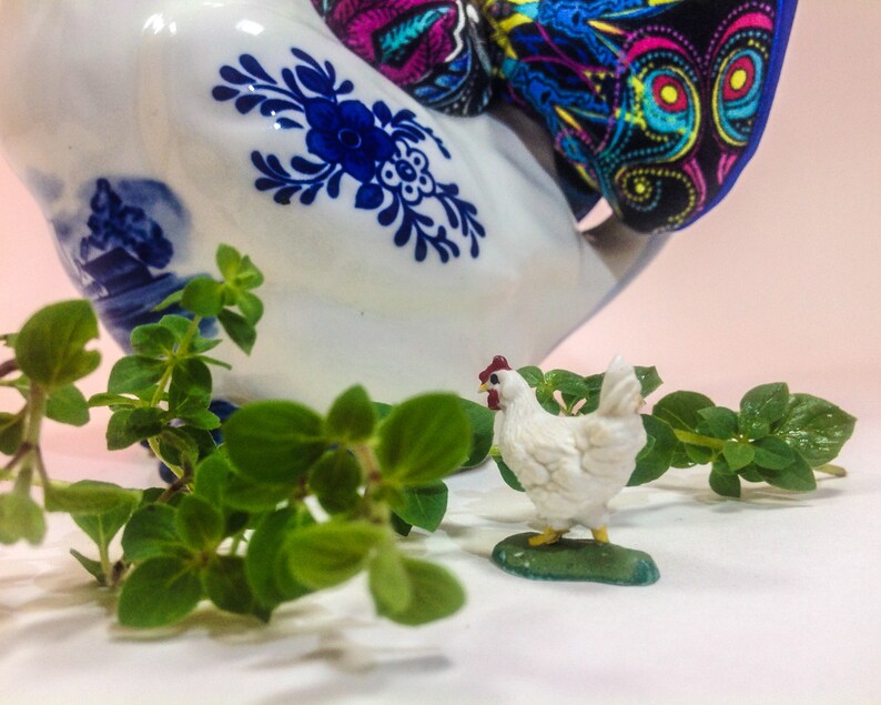 May include: A white ceramic rooster figurine with blue and white painted details, a colourful fabric scarf, and a small white chicken figurine with red accents. The figurines are surrounded by green leafy plants.