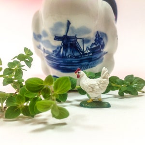 May include: A white ceramic chicken figurine with red accents stands on a green base. The figurine is in front of a blue and white ceramic jug with a windmill design. Green leafy plants are in the foreground.
