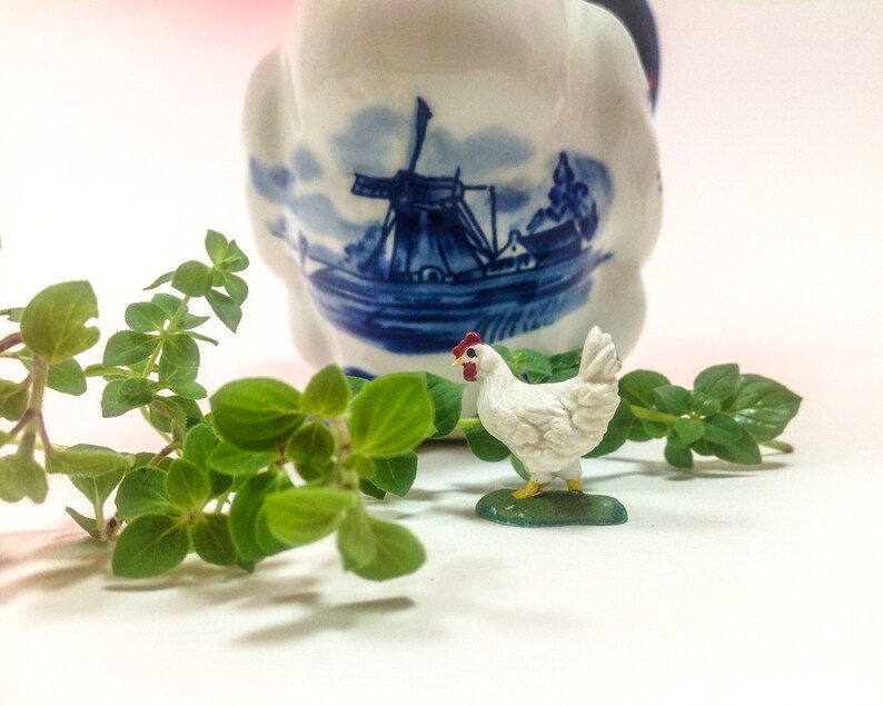 May include: A white ceramic chicken figurine with red accents stands on a green base. The figurine is in front of a blue and white ceramic jug with a windmill design. Green leafy plants are in the foreground.
