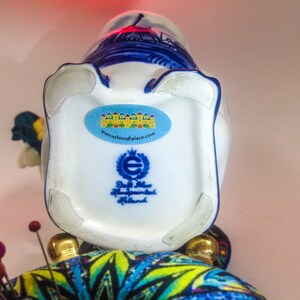 May include: A white ceramic vase with blue and white Delftware designs. The vase is decorated with a windmill and other Dutch scenes. The bottom of the vase has a sticker that says "PincushionPalace.com" and a label that says "Delft Blue Handpainted Holland".