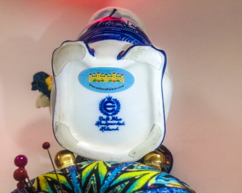 May include: A white ceramic vase with blue and white Delftware designs. The vase is decorated with a windmill and other Dutch scenes. The bottom of the vase has a sticker that says "PincushionPalace.com" and a label that says "Delft Blue Handpainted Holland".