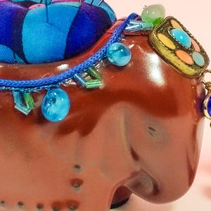 May include: A brown ceramic elephant pincushion with a blue and purple fabric pincushion on top. The elephant is decorated with blue and green beads, a gold-coloured metal ornament, and a blue gemstone pendant.