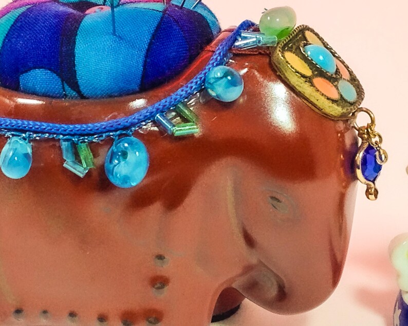 May include: A brown ceramic elephant pincushion with a blue and purple fabric pincushion on top. The elephant is decorated with blue and green beads, a gold-coloured metal ornament, and a blue gemstone pendant.