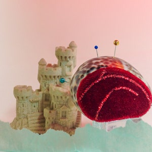 Puede incluir: Un coj&iacute;n de alfileres de terciopelo rojo con forma de concha marina con tres alfileres, situado junto a un castillo de arena en miniatura hecho de yeso beige.