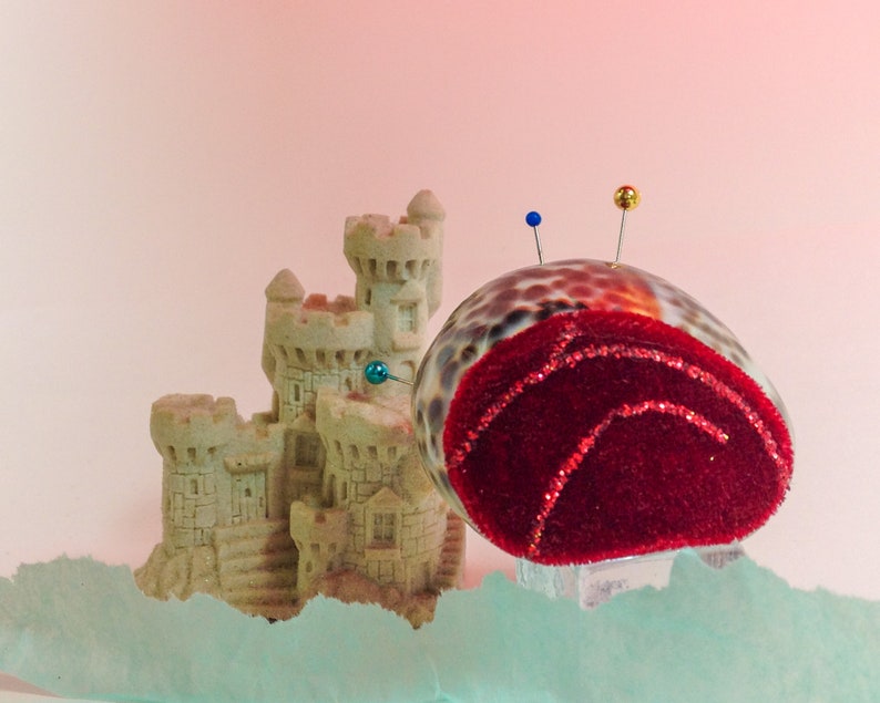 Puede incluir: Un coj&iacute;n de alfileres de terciopelo rojo con forma de concha marina con tres alfileres, situado junto a un castillo de arena en miniatura hecho de yeso beige.