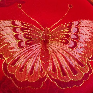 Puede incluir: Un cojín de tela roja con un diseño de mariposa bordada en oro y rosa.