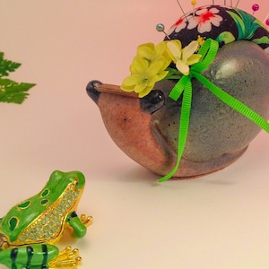 Könnte beinhalten: Ein Keramik-Nadelkissen in Form einer Schnecke mit einem grünen Band und gelben Blumen. Die Schnecke ist mit einem schwarz-weißen Blumenstoff verziert und hat eine grüne Froschfigur davor.
