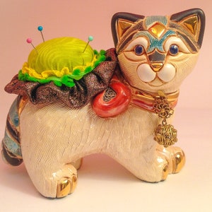 May include: A white ceramic cat figurine with a green pincushion on its back. The cat has blue eyes, a gold collar with a dangling charm, and a colorful striped tail.