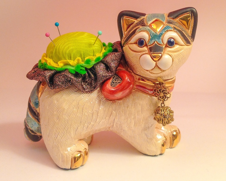 May include: A white ceramic cat figurine with a green pincushion on its back. The cat has blue eyes, a gold collar with a dangling charm, and a colorful striped tail.