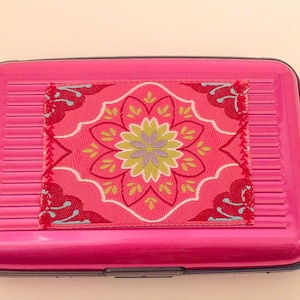 May include: A pink metal card holder with a colorful floral design on the front.