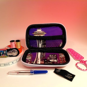 May include: A small, white and purple zippered case with a Seattle skyline and Space Needle design. The case is open and contains a variety of sewing supplies, including thread, needles, scissors, and a tape measure.