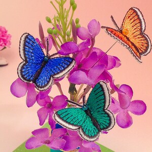 May include: A bouquet of purple flowers with three embroidered butterflies. One butterfly is blue, one is green, and one is orange.