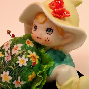 Puede incluir: Una figurita de porcelana de una niña que lleva un sombrero amarillo con un lazo rojo y un vestido verde. Está sosteniendo un cojín de alfileres verde con flores blancas y algunos alfileres.