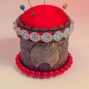 May include: A red pincushion with a silver and brown trim, surrounded by a beaded border of red beads. The pincushion is topped with three pins, one blue, one green, and one gold.