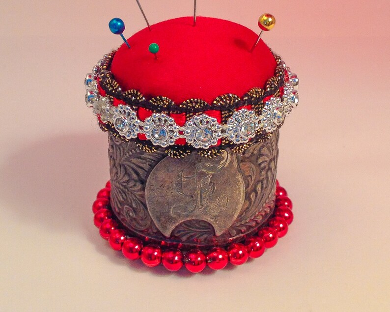 May include: A red pincushion with a silver and brown trim, surrounded by a beaded border of red beads. The pincushion is topped with three pins, one blue, one green, and one gold.