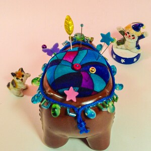 May include: A brown ceramic pincushion with a colourful fabric top and a beaded trim. The pincushion is decorated with a variety of pins, beads, and a small ceramic clown figurine.