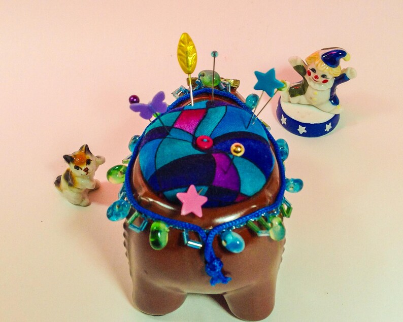 May include: A brown ceramic pincushion with a colourful fabric top and a beaded trim. The pincushion is decorated with a variety of pins, beads, and a small ceramic clown figurine.