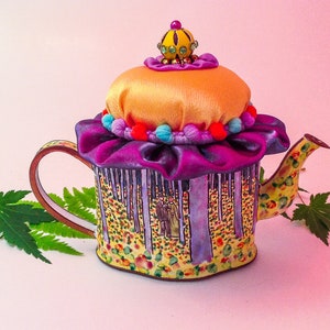 May include: A whimsical teapot with a purple and yellow fabric lid, decorated with a crown and colorful beads. The teapot is painted with a forest scene and has a yellow and white floral pattern.