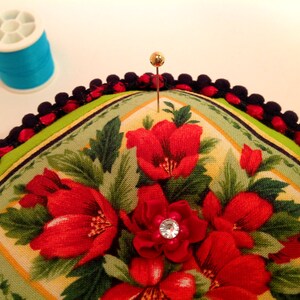May include: A green and yellow floral pincushion with a black and red trim. A gold pin is stuck in the center of the pincushion.