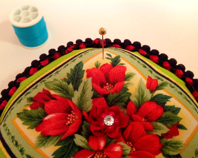 May include: A green and yellow floral pincushion with a black and red trim. A gold pin is stuck in the center of the pincushion.