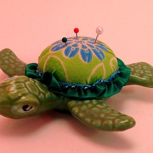 以下が含まれることがあります： 青と緑の花柄の布地で覆われた緑色のセラミック製の亀の針山。針山はベースの周りに緑色のフリルが付いており、カラフルなピンで飾られています。