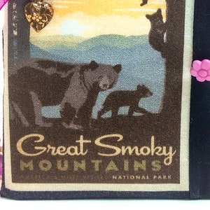May include: A black fabric pouch with a zipper closure. The front of the pouch features a vintage-style illustration of bears in the Great Smoky Mountains National Park. The pouch has a pink flower button and a gold heart charm.
