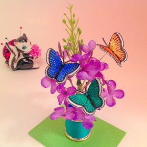 May include: A small floral arrangement with purple flowers and three embroidered butterflies: blue, green, and orange. The arrangement is in a small teal blue spool.