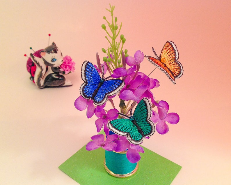 May include: A small floral arrangement with purple flowers and three embroidered butterflies: blue, green, and orange. The arrangement is in a small teal blue spool.