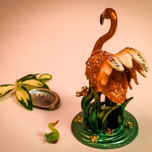 May include: A decorative figurine of a flamingo with a jeweled design, standing on a green base with a small duck figurine in the foreground.