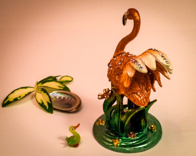 May include: A decorative figurine of a flamingo with a jeweled design, standing on a green base with a small duck figurine in the foreground.