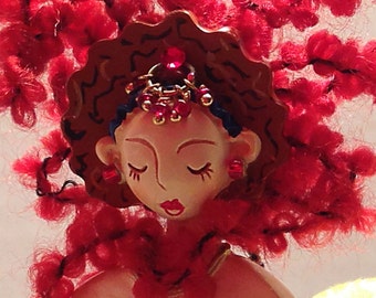 Puede incluir: Una muñeca de pelo rojo con los ojos cerrados y una corona roja y dorada. La muñeca lleva un vestido rojo y tiene un fondo rojo.