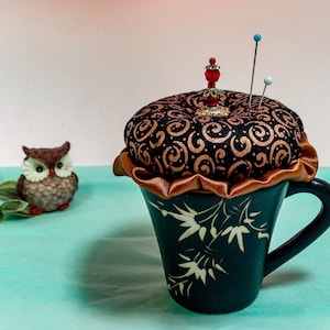 Pode incluir: Uma chávena de chá de cerâmica preta com um desenho floral branco e um almofada de alfinetes de tecido castanho com detalhes dourados no topo. A almofada de alfinetes tem um ornamento de contas vermelhas e dois alfinetes.