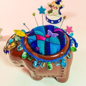 May include: A brown ceramic elephant pincushion with a colourful fabric top and blue and green beaded trim. The pincushion has a variety of colourful pins and a yellow feather sticking out of it.