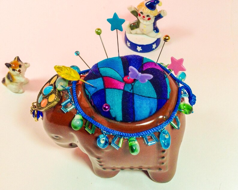 May include: A brown ceramic elephant pincushion with a colourful fabric top and blue and green beaded trim. The pincushion has a variety of colourful pins and a yellow feather sticking out of it.