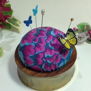May include: A round pincushion with a pink and blue floral pattern, sitting on a wooden base with a brass finish. The pincushion has several pins with blue and yellow butterfly embellishments.