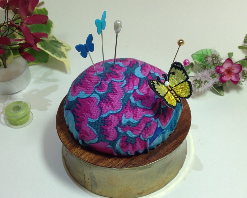 May include: A round pincushion with a pink and blue floral pattern, sitting on a wooden base with a brass finish. The pincushion has several pins with blue and yellow butterfly embellishments.