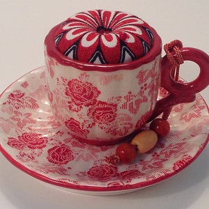 May include: A white ceramic teacup and saucer with a red floral pattern. The teacup has a red fabric pincushion with a white and black floral design inside. A red and wooden beaded string hangs from the teacup handle.