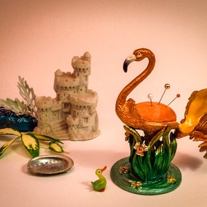 May include: A gold flamingo pincushion with an open beak and a red, white, and orange pincushion inside. The flamingo is standing on a green base with a floral design. There is a small green duck figurine and a seashell on the table.