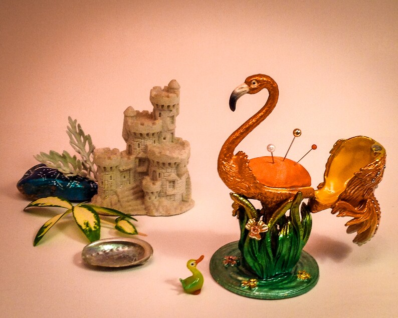 May include: A gold flamingo pincushion with an open beak and a red, white, and orange pincushion inside. The flamingo is standing on a green base with a floral design. There is a small green duck figurine and a seashell on the table.