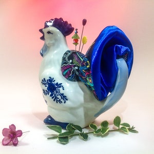 May include: A white ceramic chicken pincushion with blue and white floral designs. The chicken has a blue satin bow on its tail and colourful pins sticking out of its back.