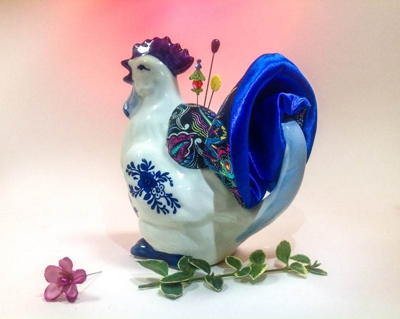 May include: A white ceramic chicken pincushion with blue and white floral designs. The chicken has a blue satin bow on its tail and colourful pins sticking out of its back.