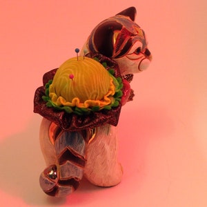 May include: A ceramic cat figurine with a colorful pincushion on its back. The pincushion is yellow with green and red accents. The cat is white with black and blue markings.