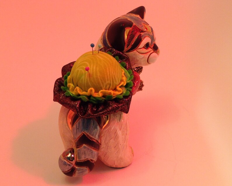 May include: A ceramic cat figurine with a colorful pincushion on its back. The pincushion is yellow with green and red accents. The cat is white with black and blue markings.